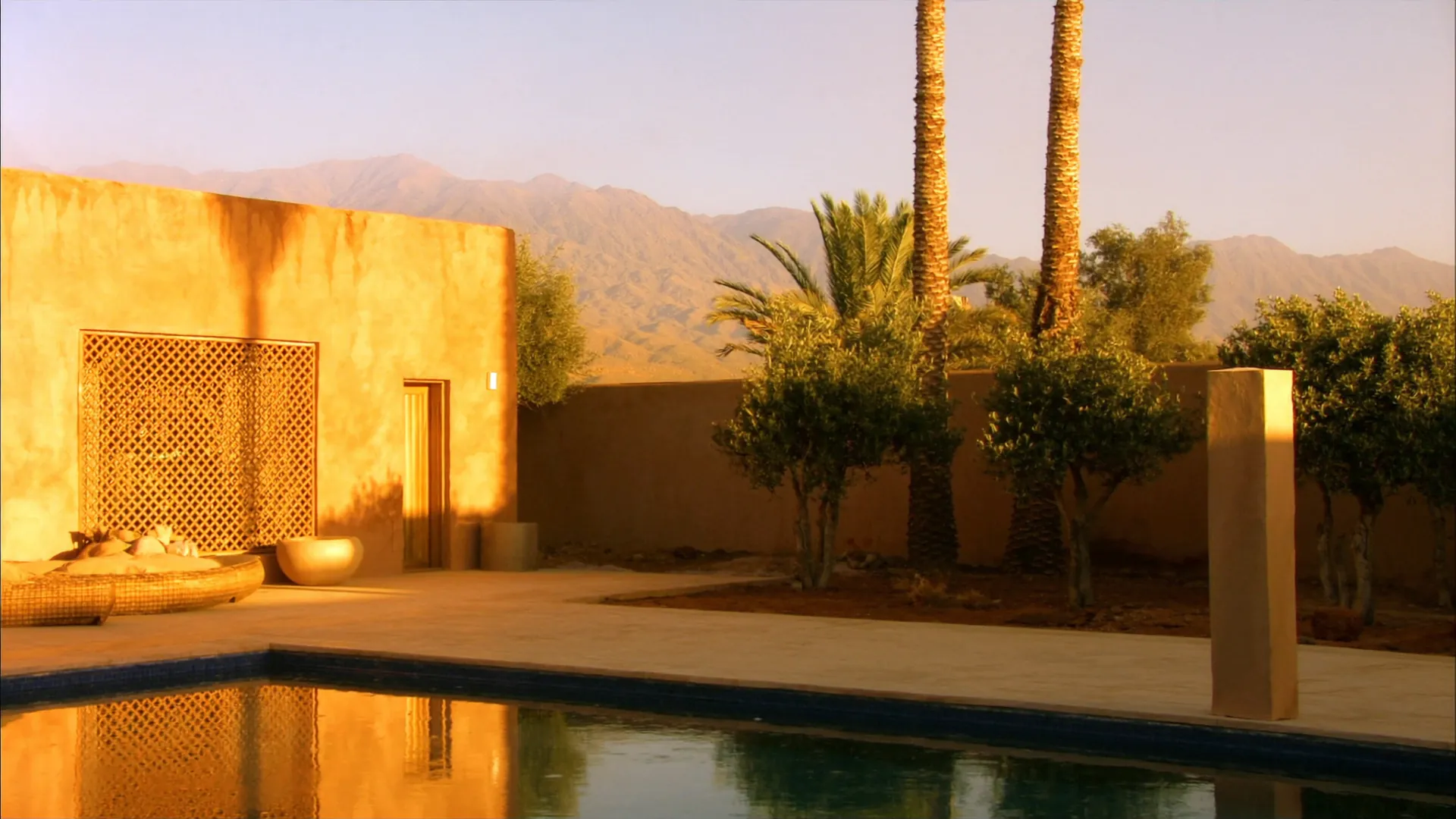 La terrasse d’une villa marrakchie au soleil couchant : bassin, oliviers, palmiers et l’Atlas au loin